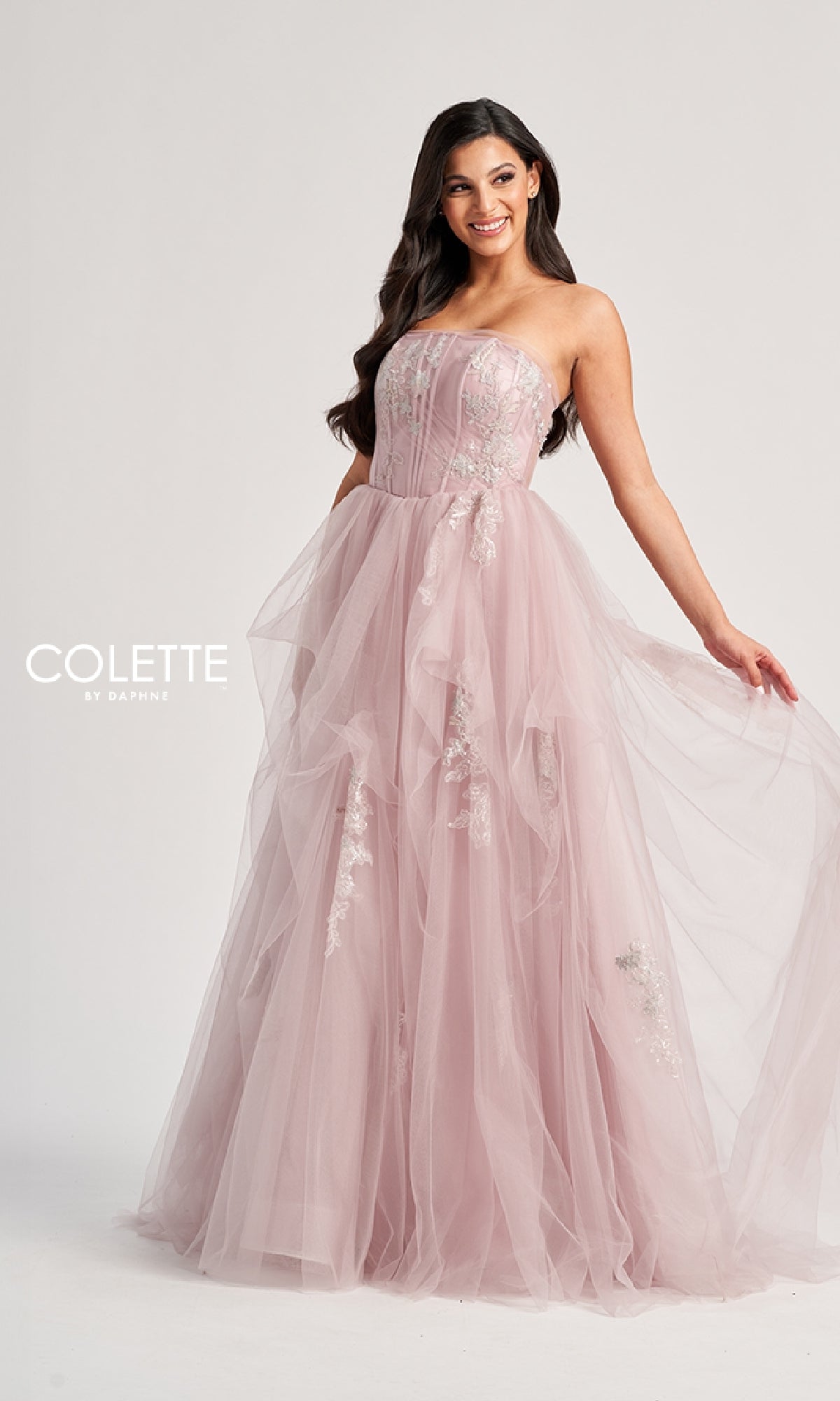 A woman smiles against a plain background, wearing the Colette CL8120 long prom dress with a strapless bodice, tulle A-line skirt, and delicate floral embellishments.